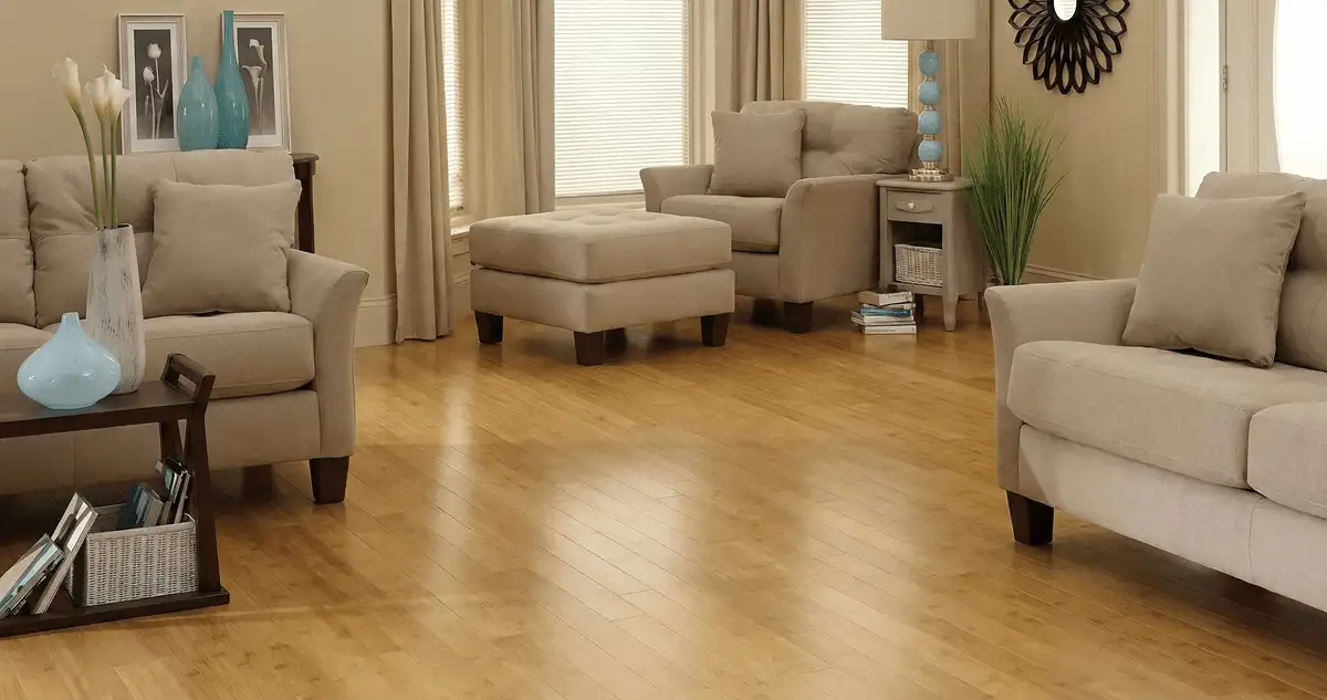 A living room with professionally installed bamboo flooring, beige sofas, a matching ottoman, a side table with a blue lamp, vases, framed pictures, and large windows with blinds and curtains in Portland, OR